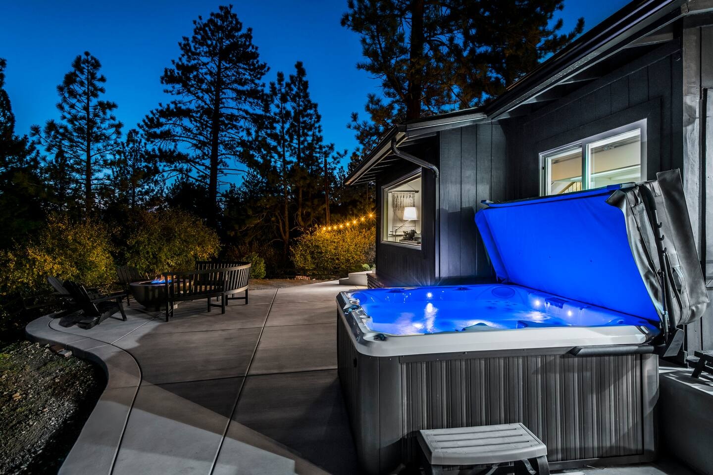 Hot tub glowing blue at dusk with fire pit and string lights in the background